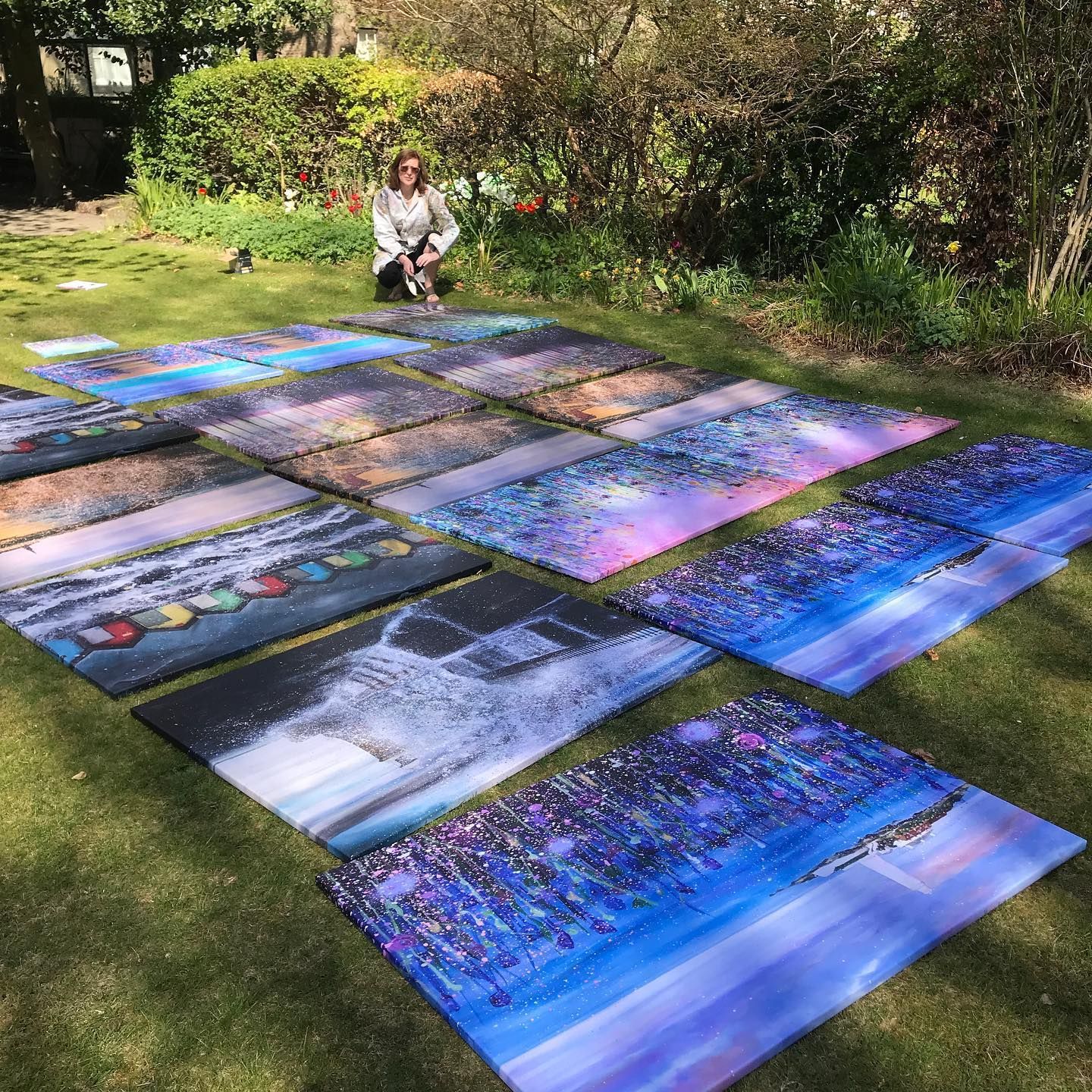 Person kneeling behind colorful landscape paintings laid out on grass in a sunny garden
