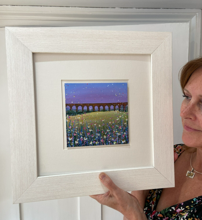 Woman holding a white-framed painting of a colorful flower field and arched bridge