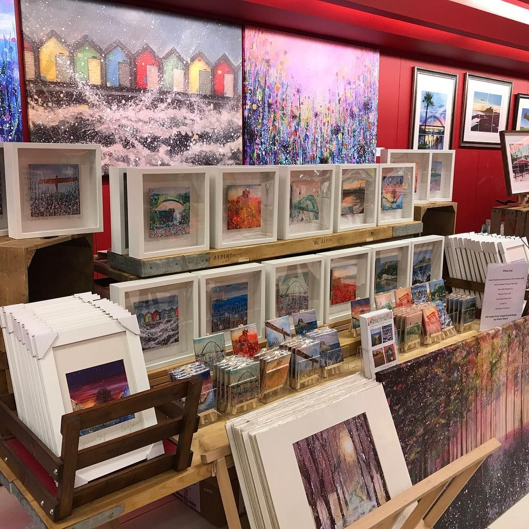 Art booth with framed paintings, prints, and cards displayed on tables against a red wall.