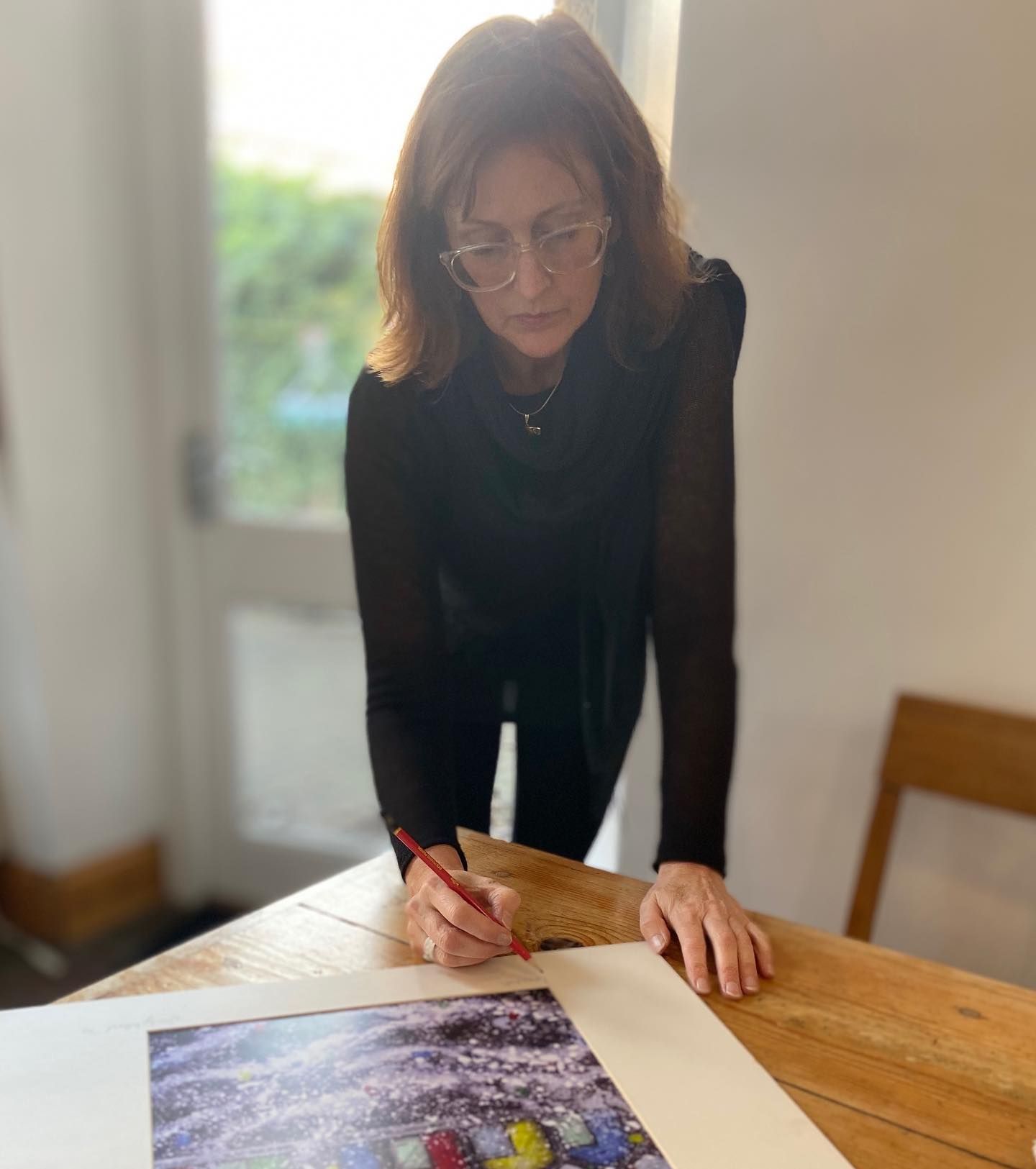 Woman leaning over a wooden table, writing on a page beside a colorful photo print.