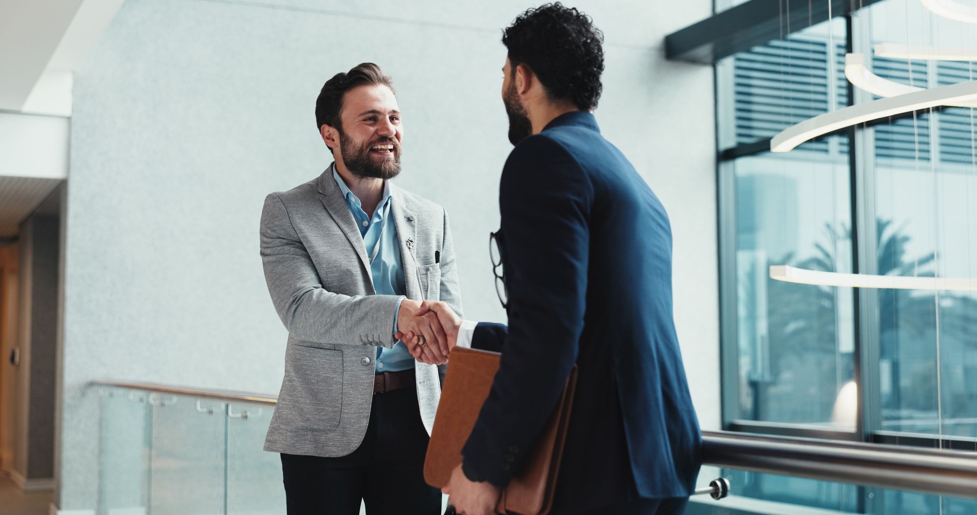 Two business people in suits shake hands while standing in a bright, modern office hallway.