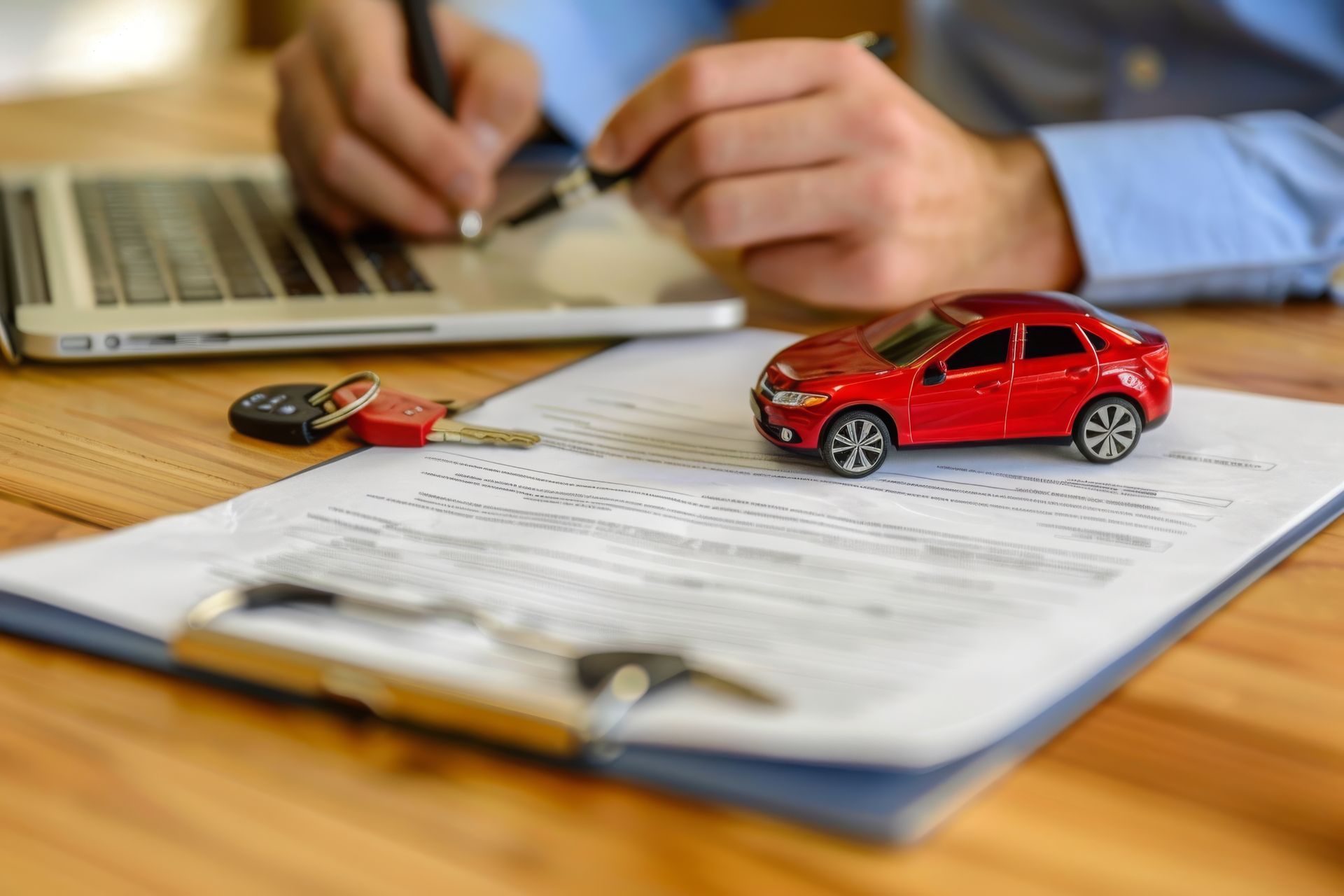Car insurance agency agent filling out a policy form near a red toy car, keys, and a silver laptop.