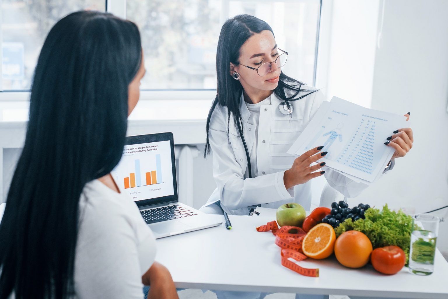 Nutritionist explaining a meal plan to a client at a desk with fruits, vegetables, and a laptop chart