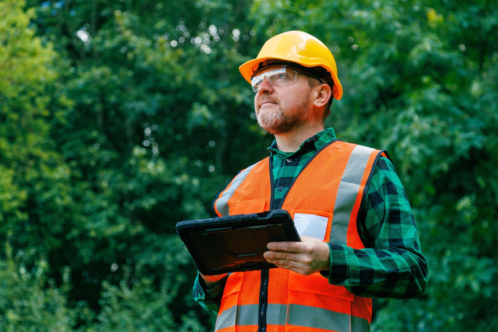 A man wearing an orange safety vest & glasses is holding a tablet, focused on his inspection.