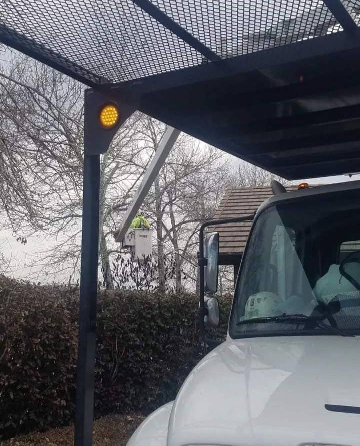 A utility truck with a yellow light on its roof frame, with a worker in a bucket lift visible in the background trees.