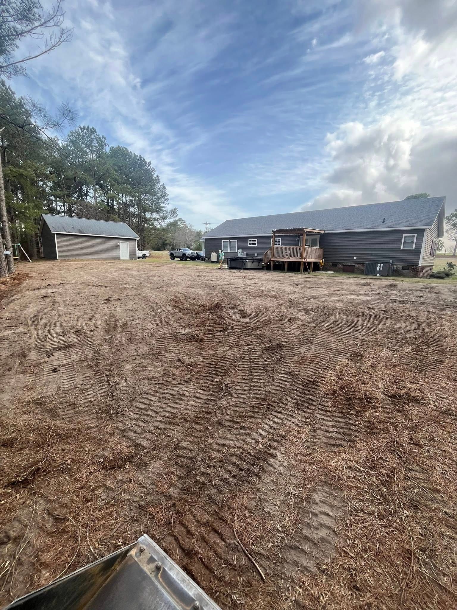 Wide gravel driveway beside dark-sided buildings, with trees and a cloudy blue sky overhead.