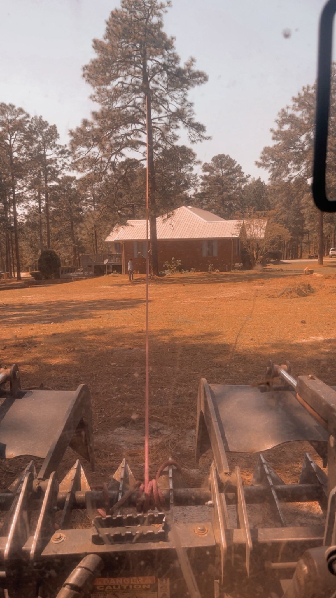 View from farm machinery toward a small house and trees in a dry, orange-brown yard
