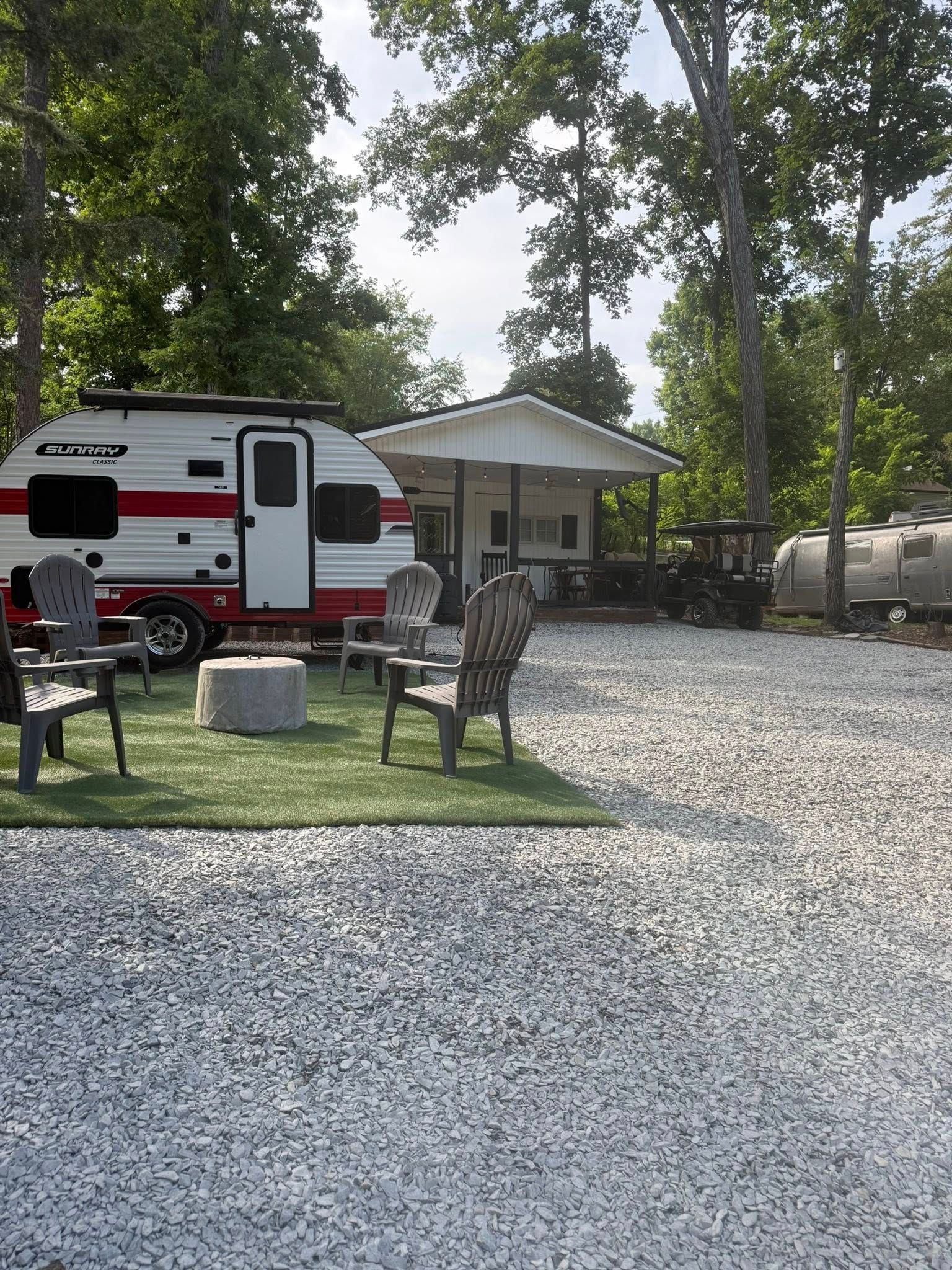 Camper parked beside a small cabin in a wooded RV campsite with gravel and patio chairs.