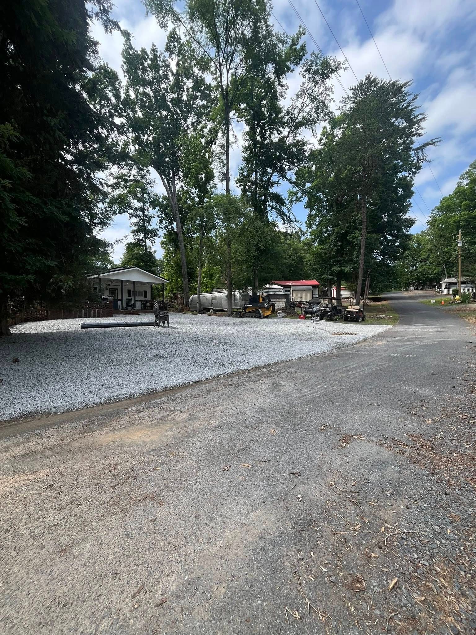 Gravel lot with trees, a white rock pile, and a small building in the background under a partly cloudy sky