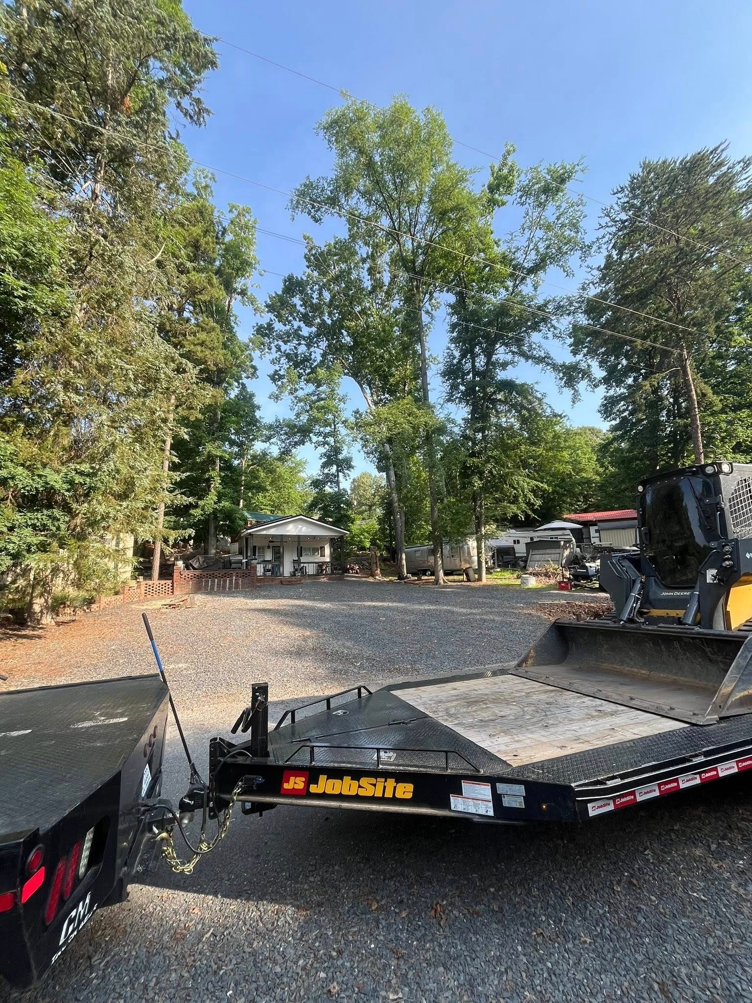 Flatbed trailer loaded with gravel, parked outdoors among trees and vehicles.