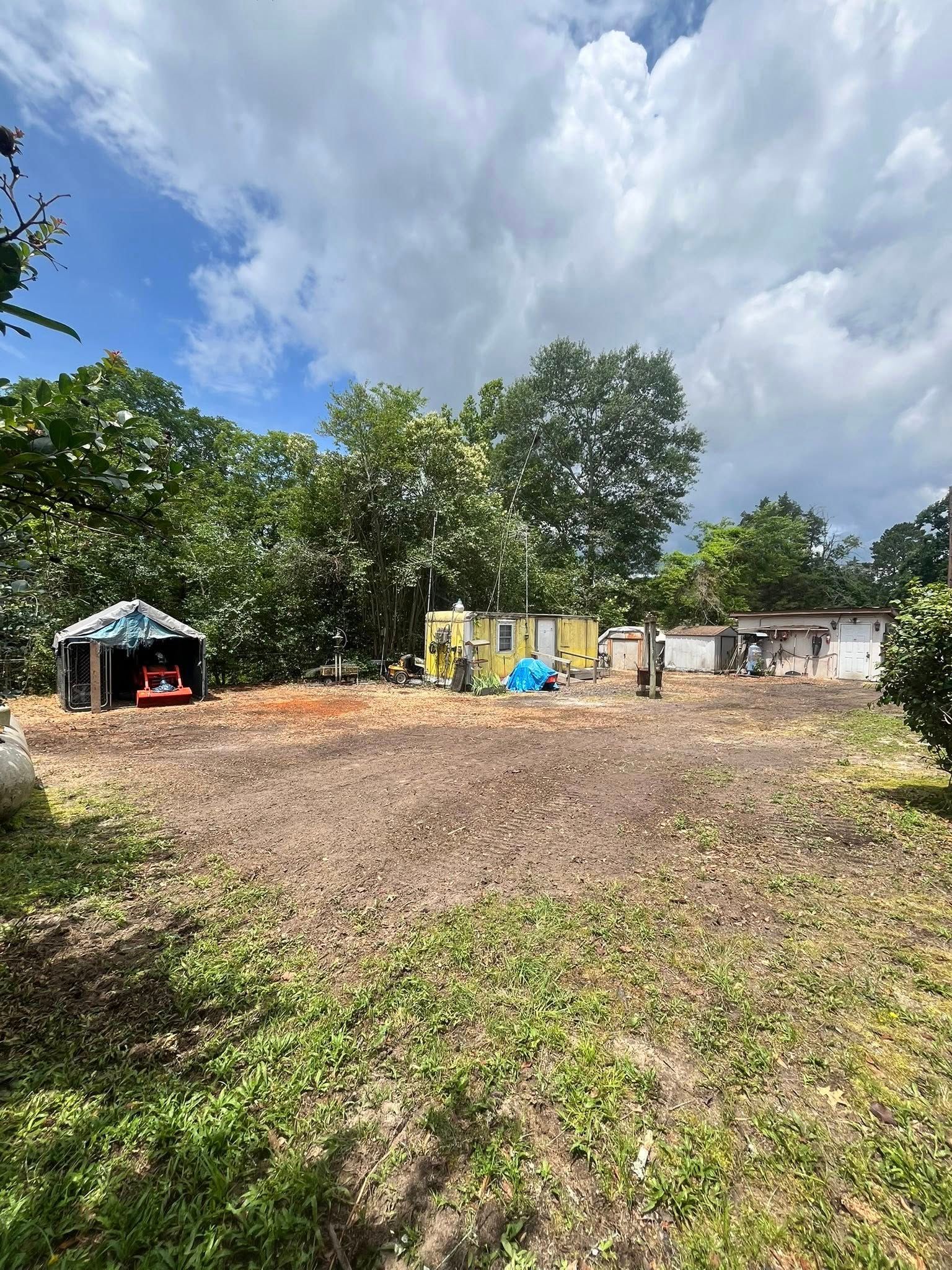 Rural open field with a small shelter, scattered items, trees, and a partly cloudy sky.