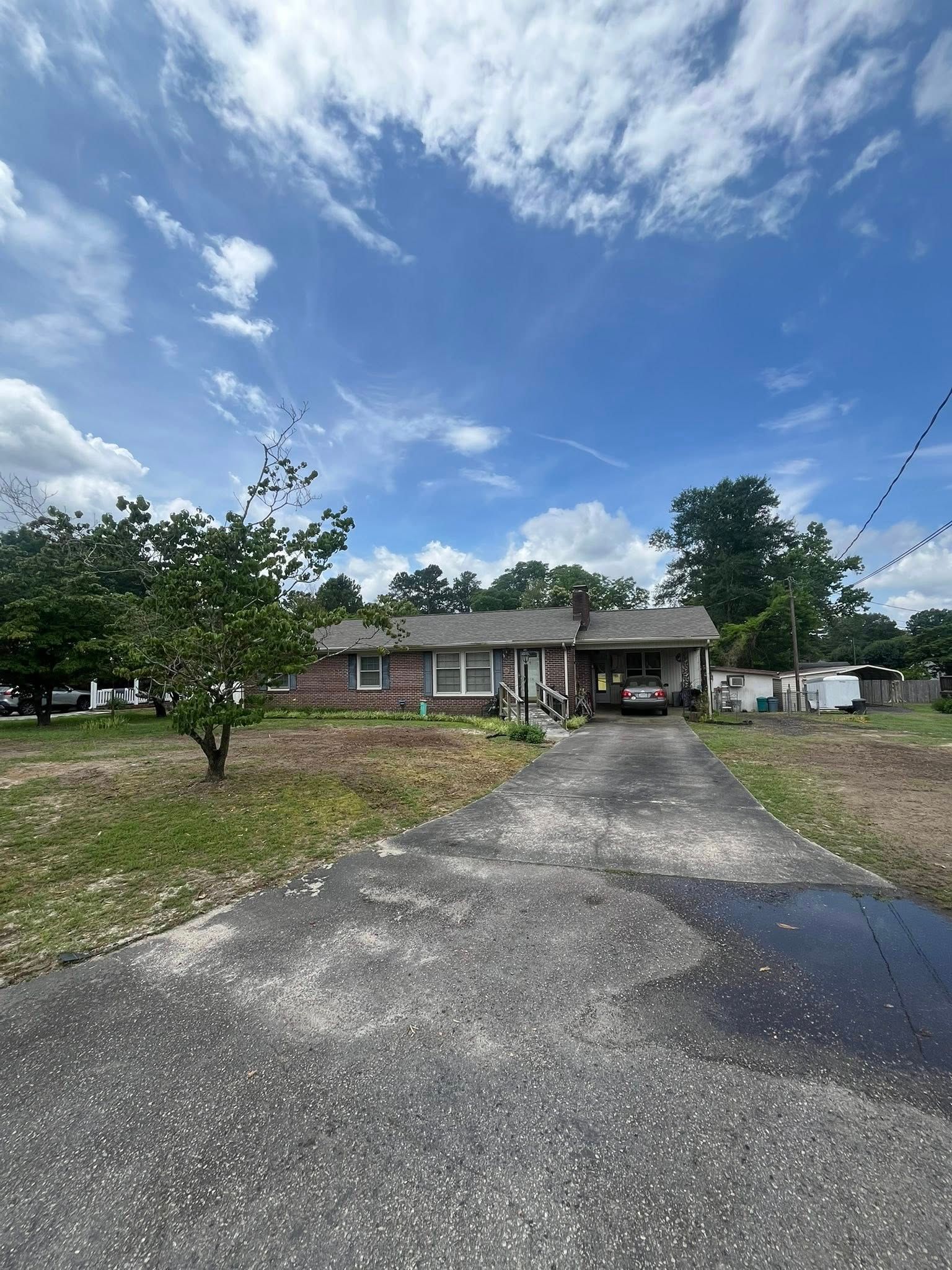 Single-story brick house with a driveway, trees, and a cloudy blue sky.
