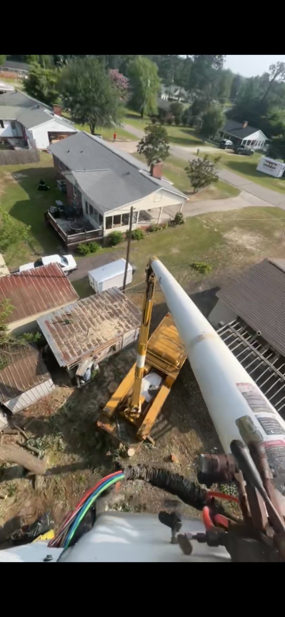 Aerial view of a construction site with a crane lifting a long white pole beside houses and trees.