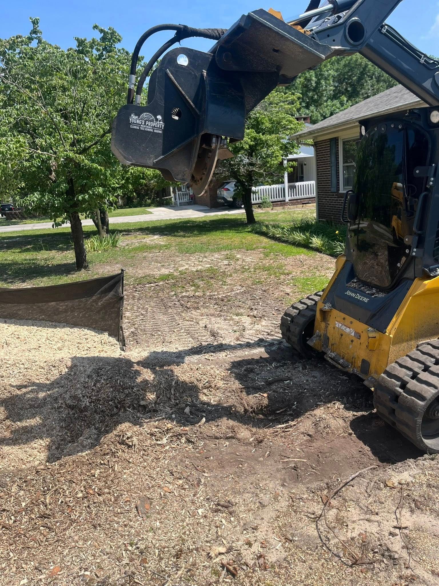 Excavator with claw lifting debris in a sunny yard beside a house and trees