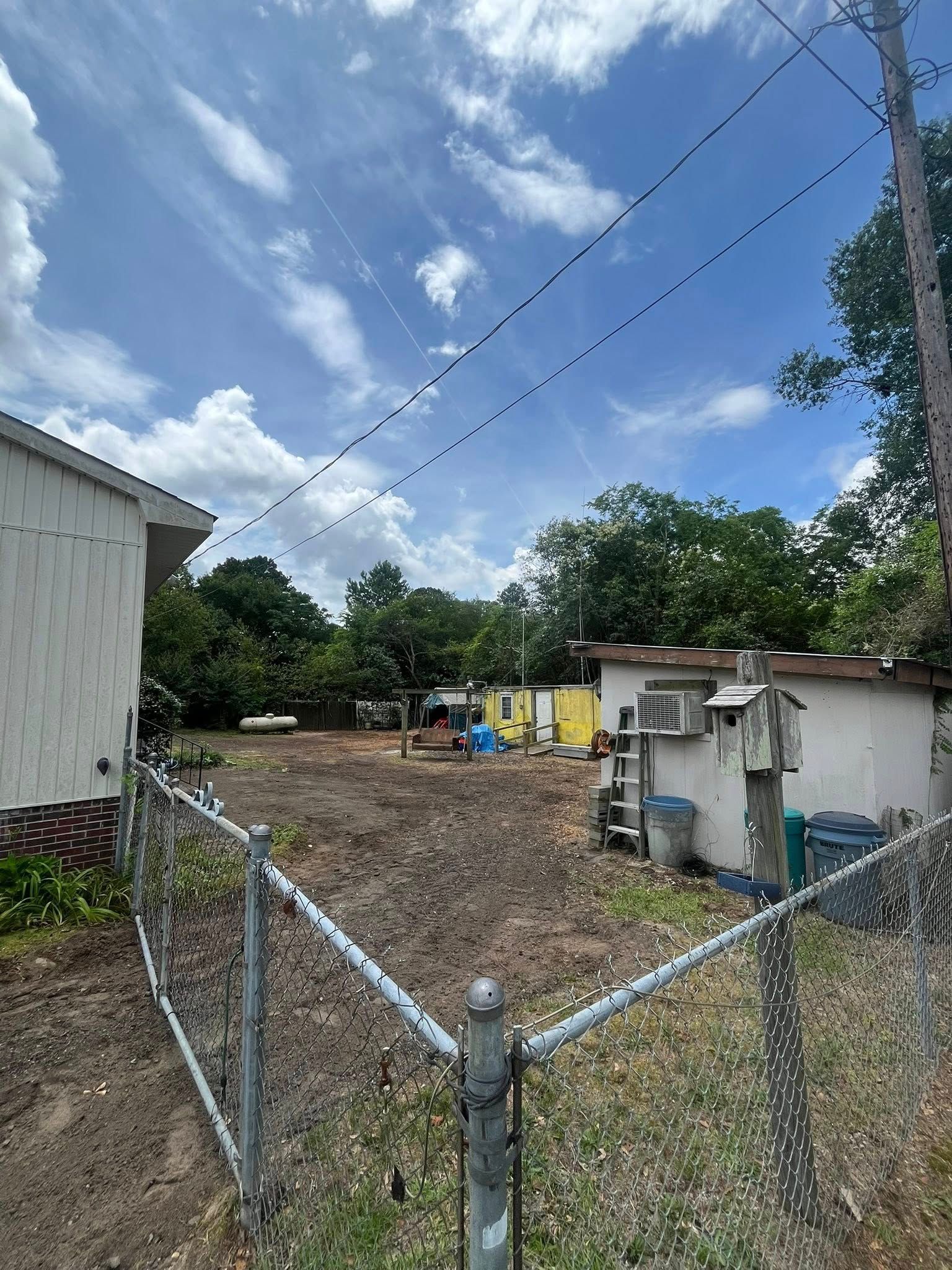 Chain-link fenced yard with a small shed, trees, and a partly cloudy sky