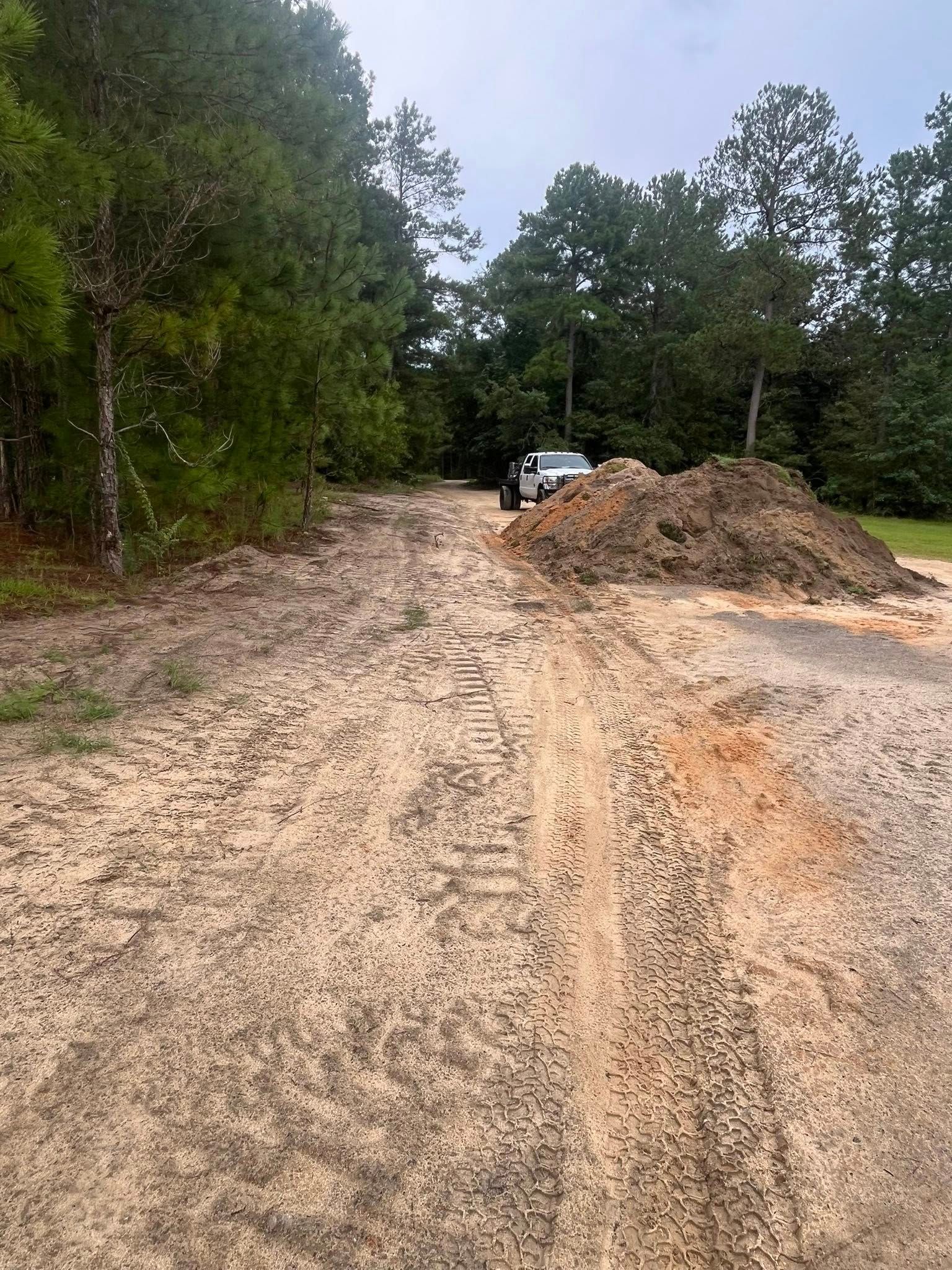 Rutted dirt road in a wooded area with a pile of sand and a white truck ahead