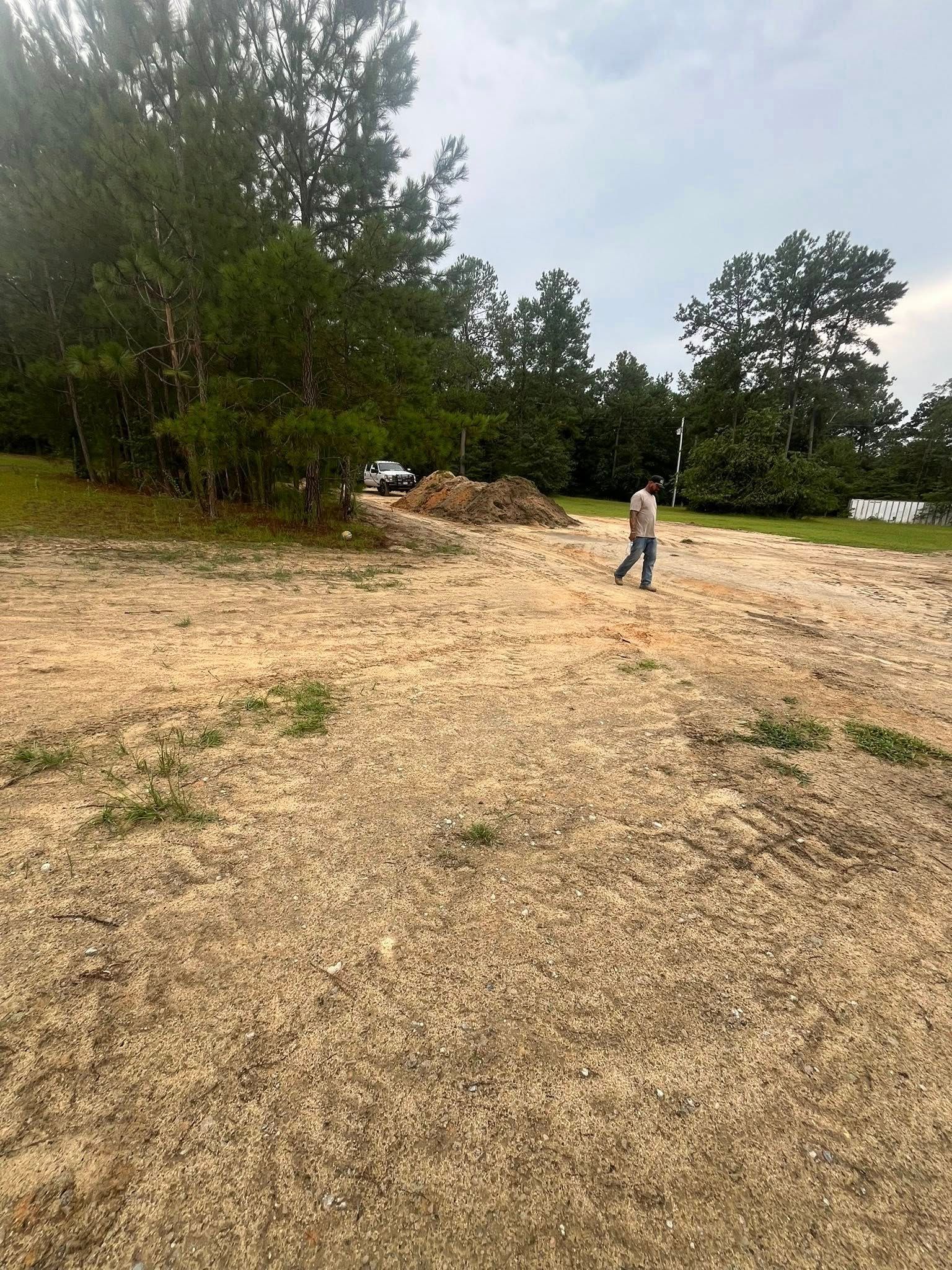 Person standing in a cleared dirt lot near trees and a pile of debris under an overcast sky
