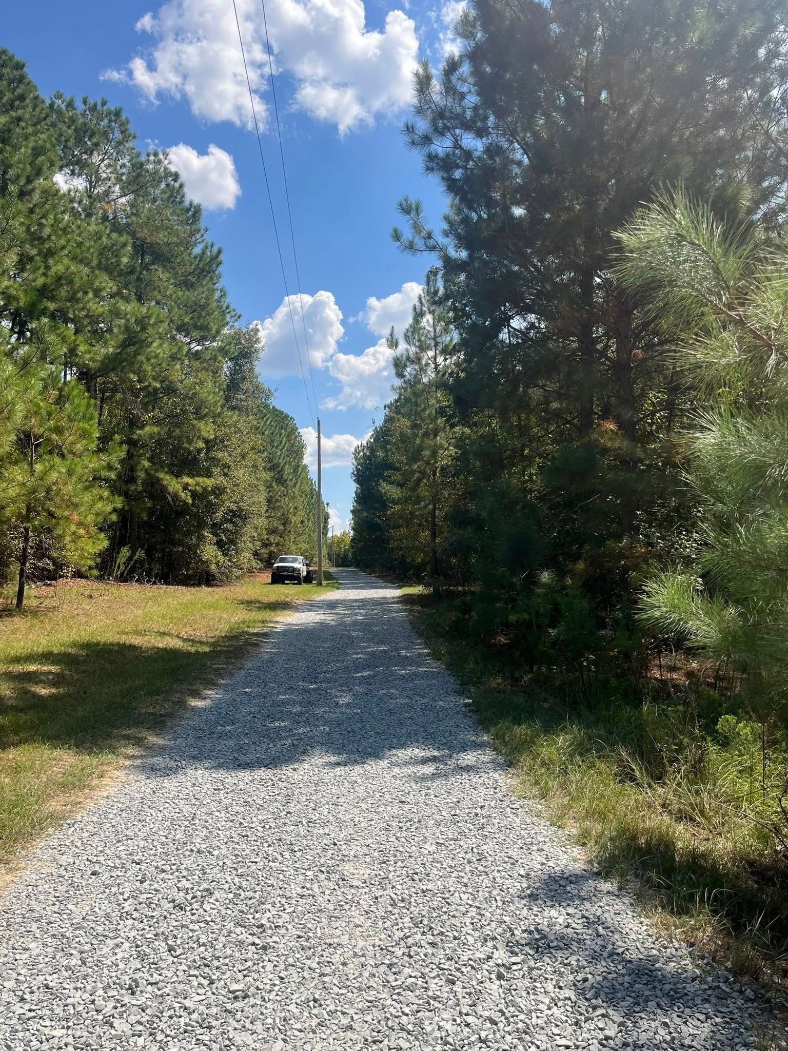 Gravel driveway lined with trees under a blue sky with clouds, leading toward a distant monument and parked car