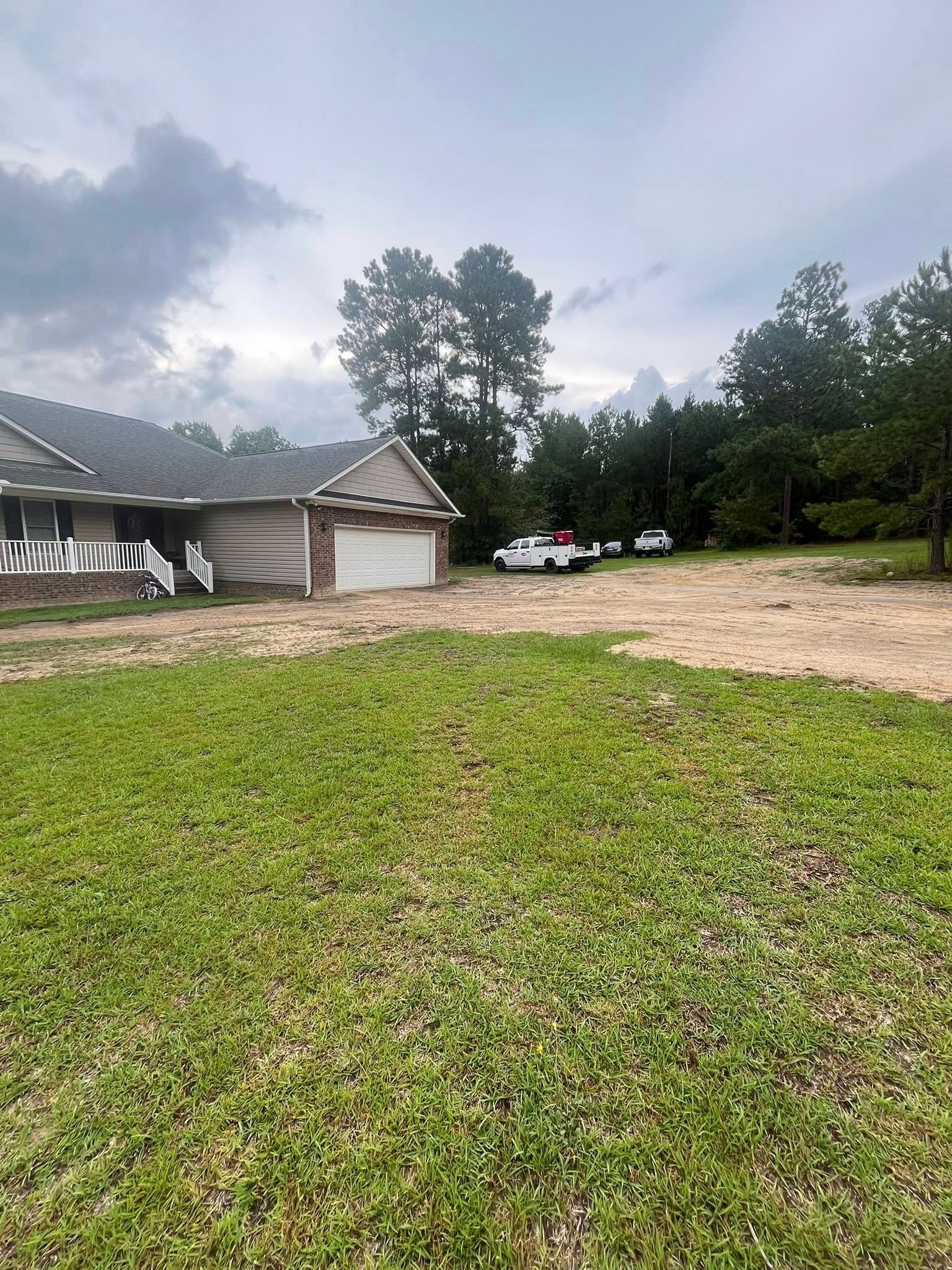 Grassy yard beside a house, with a gravel driveway, parked cars, and cloudy sky.