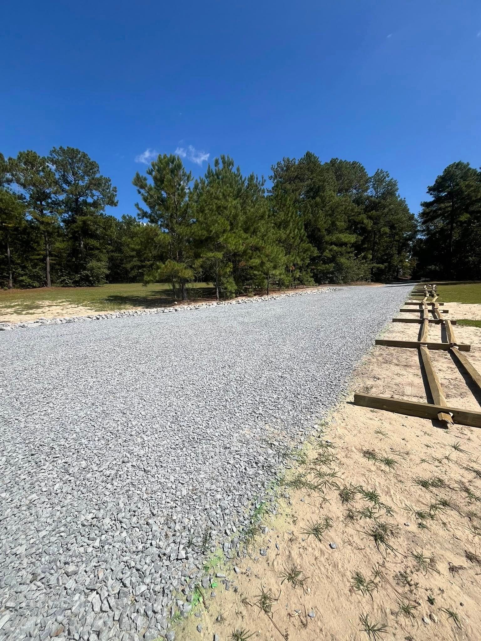 Gravel-lined path beside sandy ground and wooden posts under a blue sky, with trees in the background