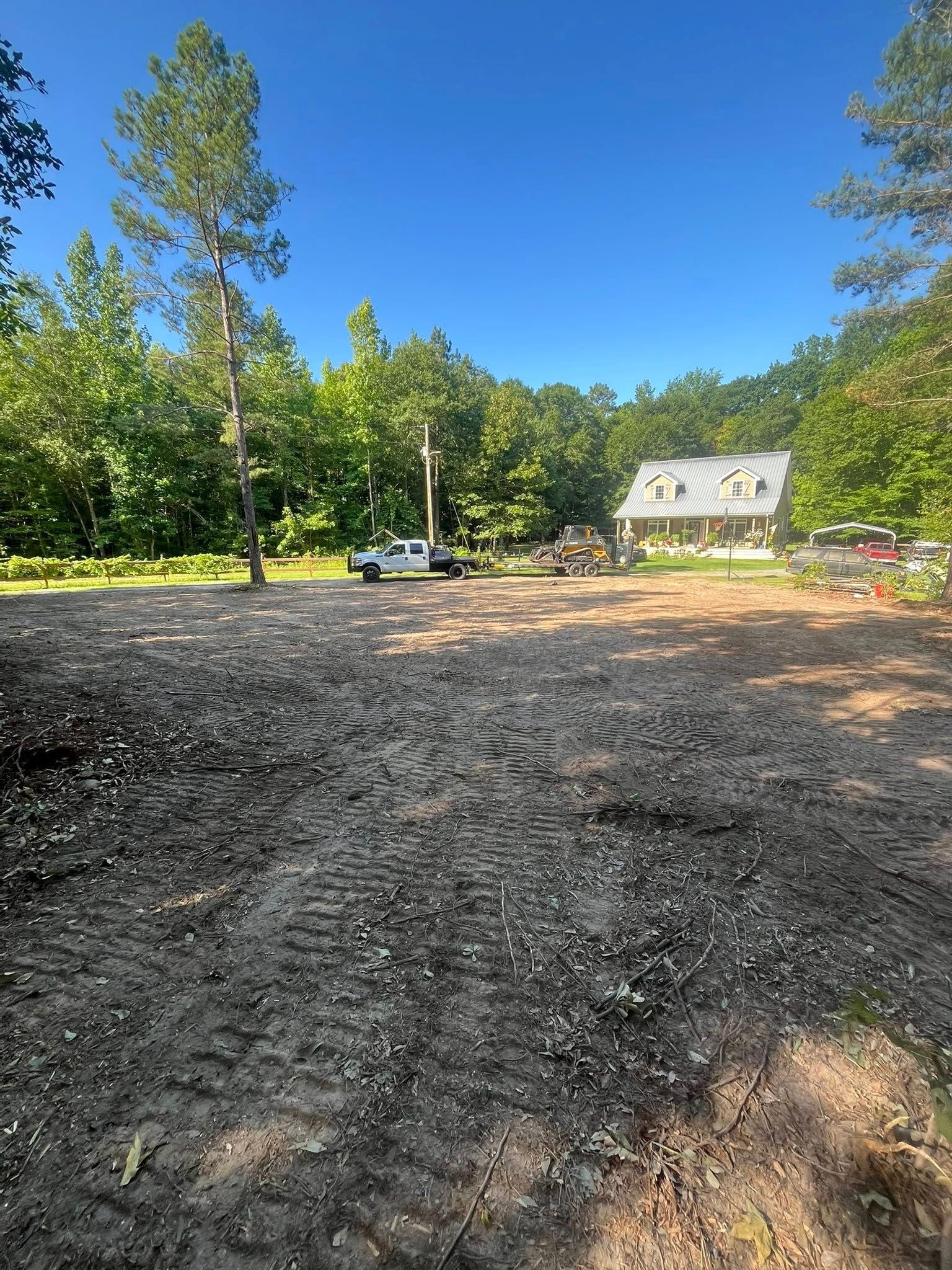 Gravel lot with trees, a parked car, and a house in the background under a clear blue sky