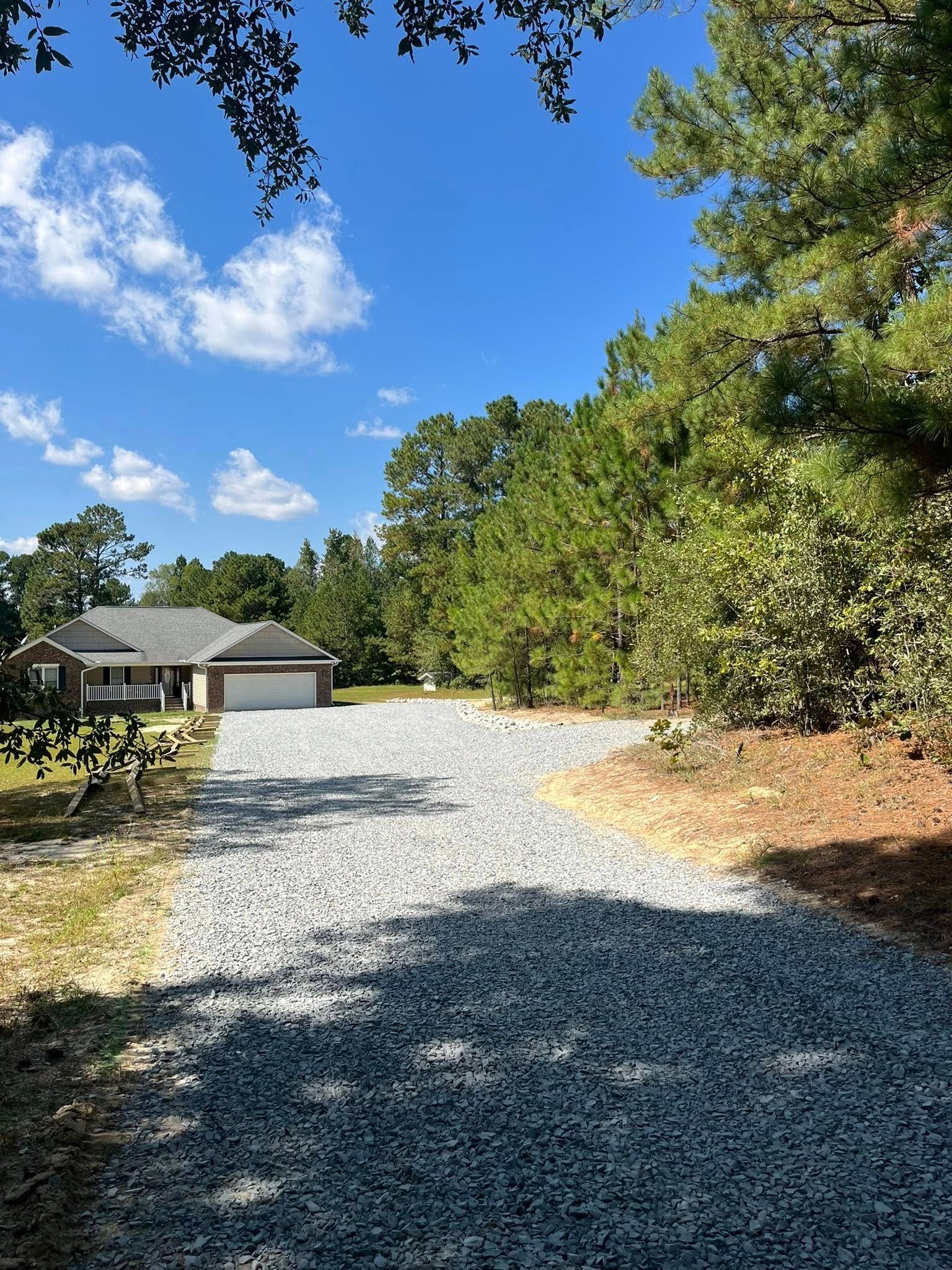 Gravel driveway leading to a house, bordered by trees under a bright blue sky.