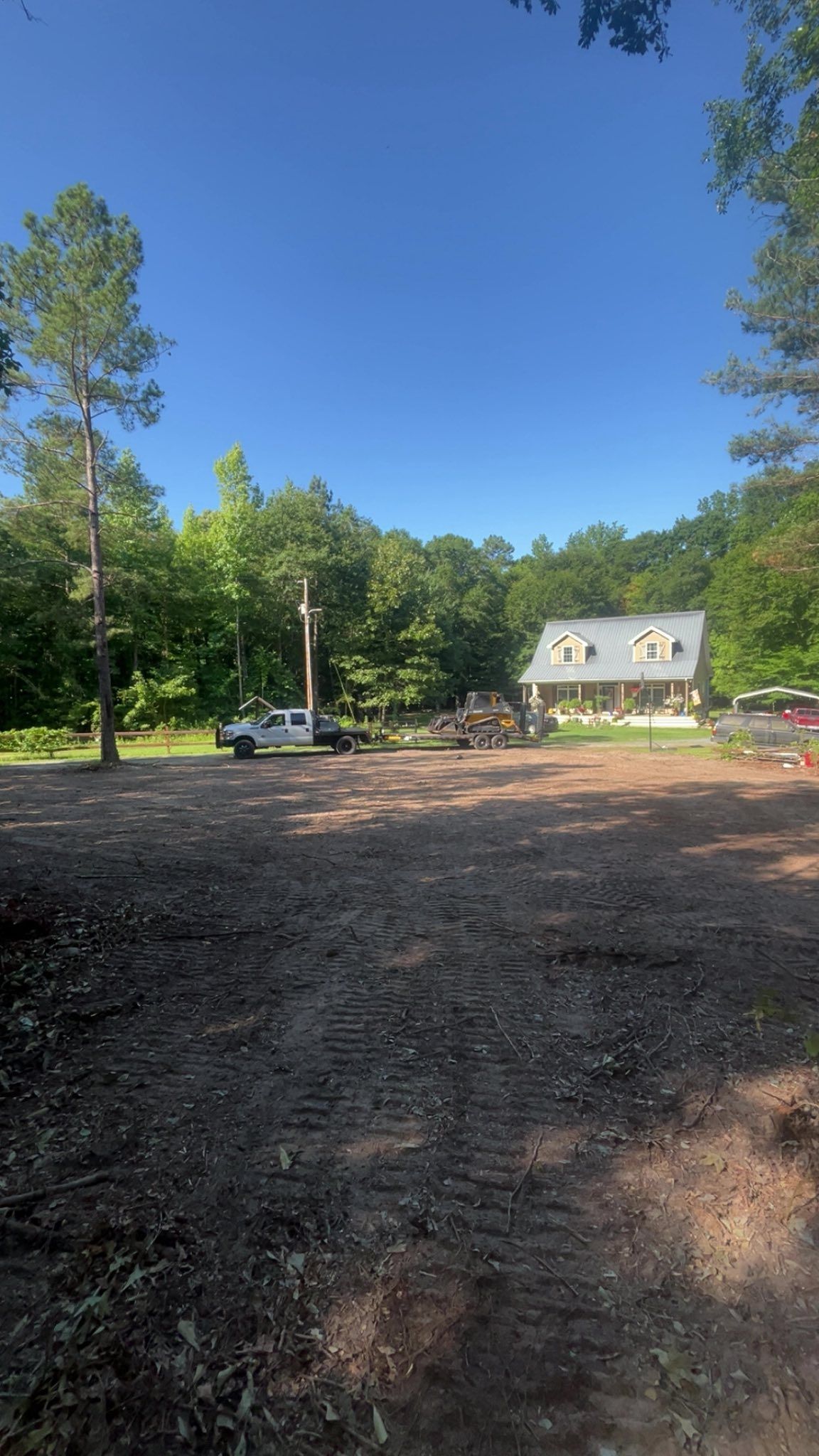Sunny wooded lot with a gravel clearing, parked cars, and a small house in the background.