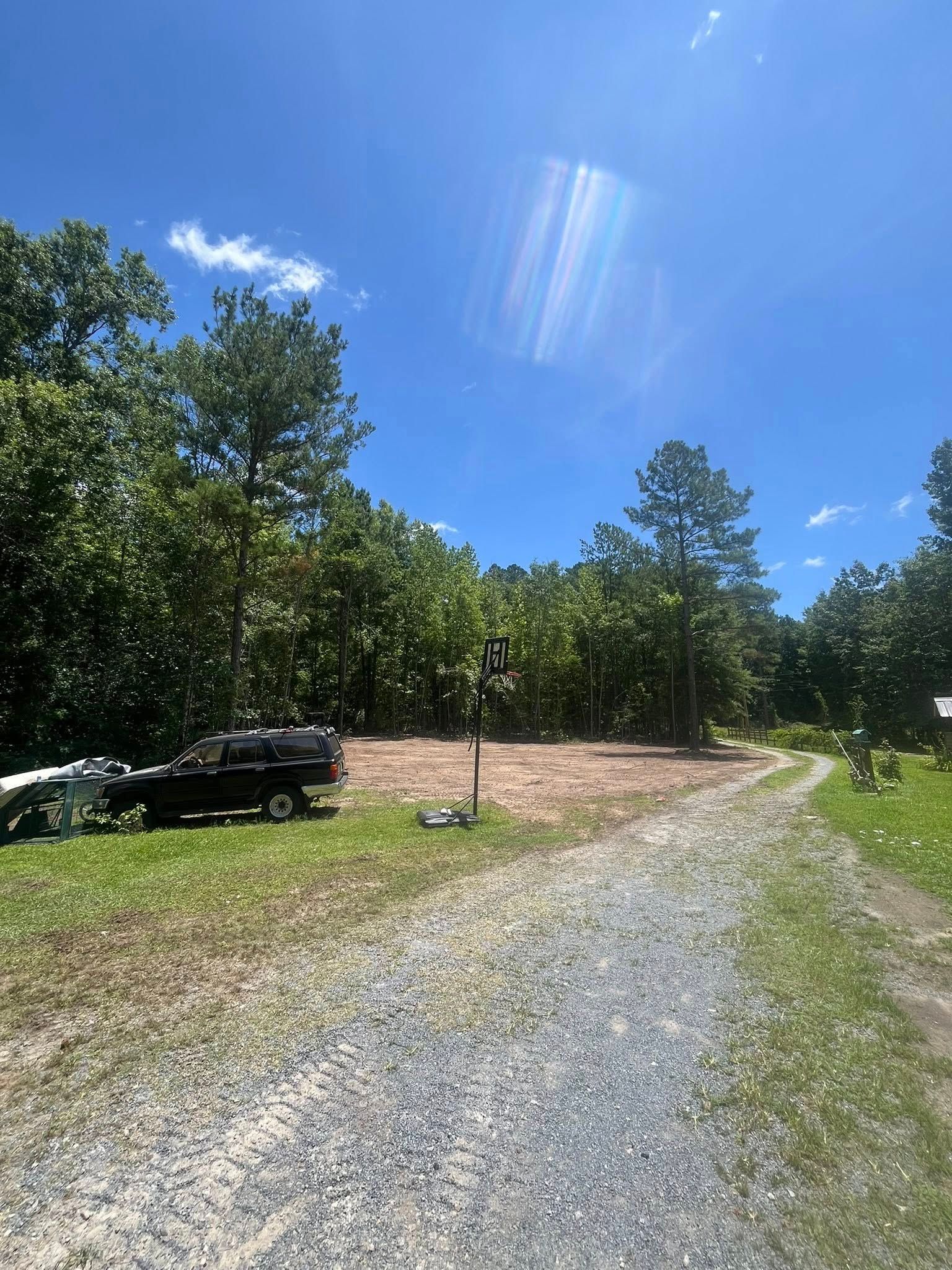 Gravel driveway curving past parked black vehicle toward wooded area under bright blue sky