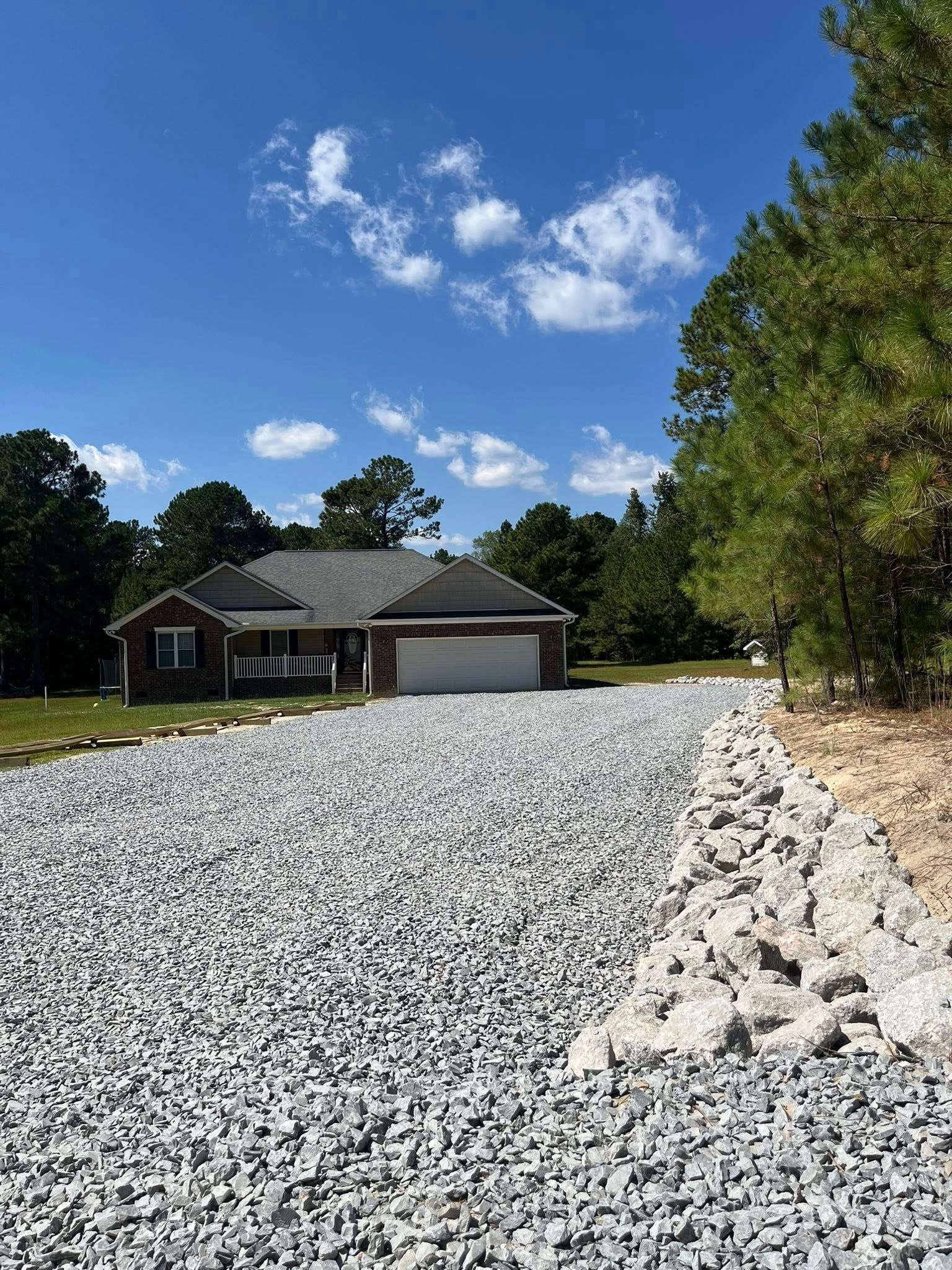 Gravel driveway leading to a gray house with a garage under a blue sky with trees.