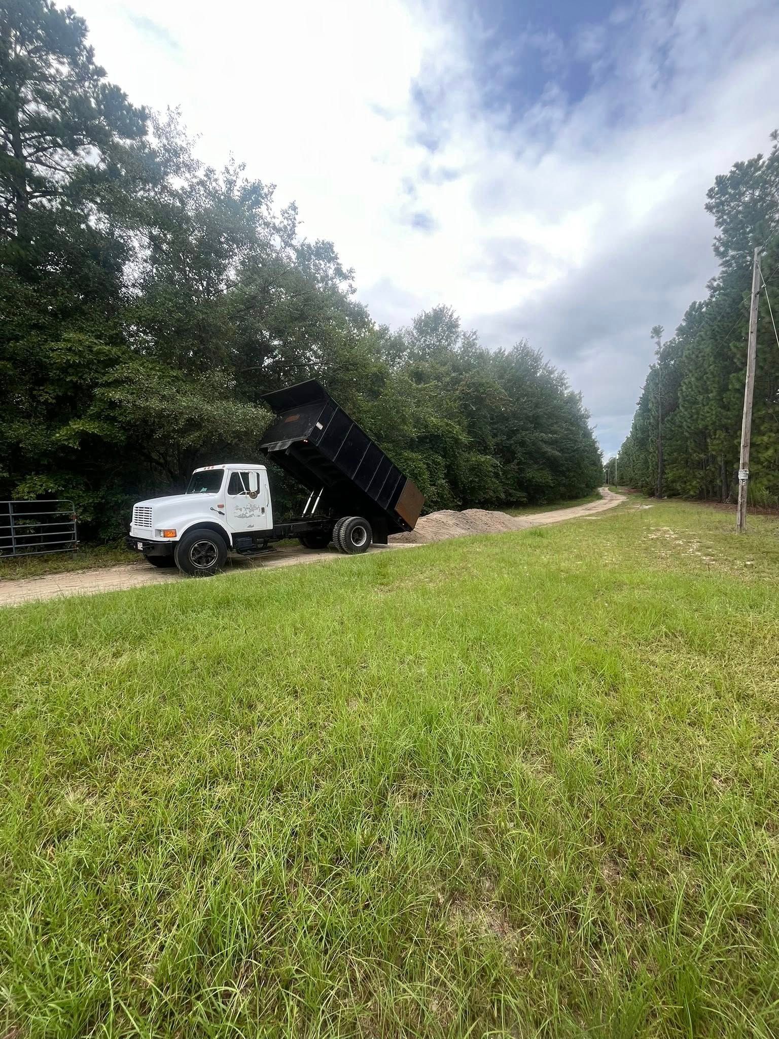 White dump truck unloading dirt beside a grassy roadside in a wooded area under cloudy skies