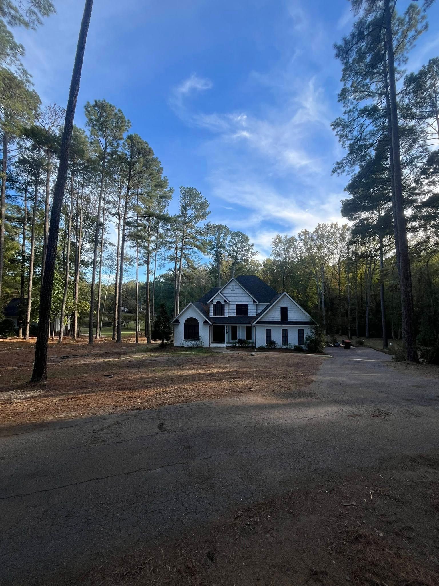 White house with dark roof among tall pine trees on a quiet gravel driveway under a blue sky.