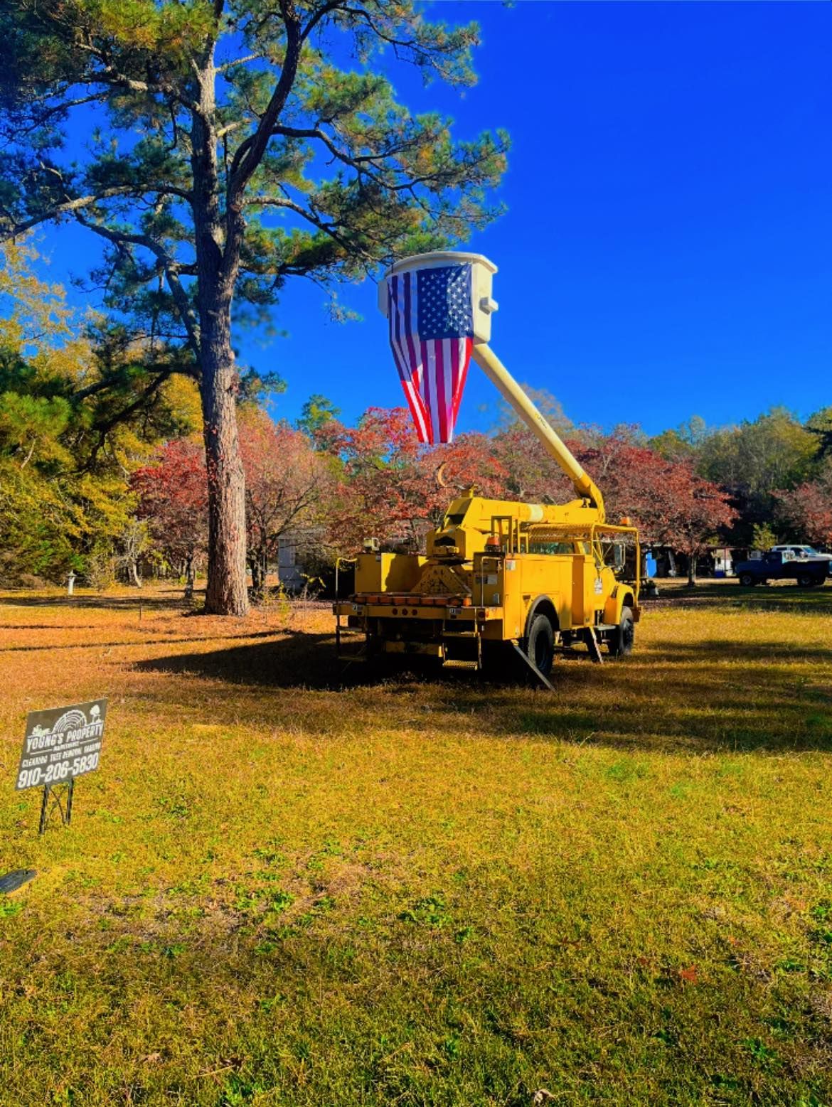 Yellow cherry picker truck raising a large American flag in a grassy park under a bright blue sky