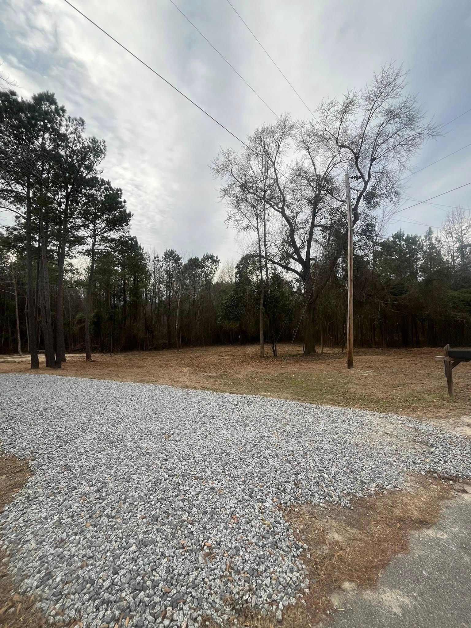 Wide gravel clearing beside trees under a cloudy sky, with power lines overhead and a utility pole.