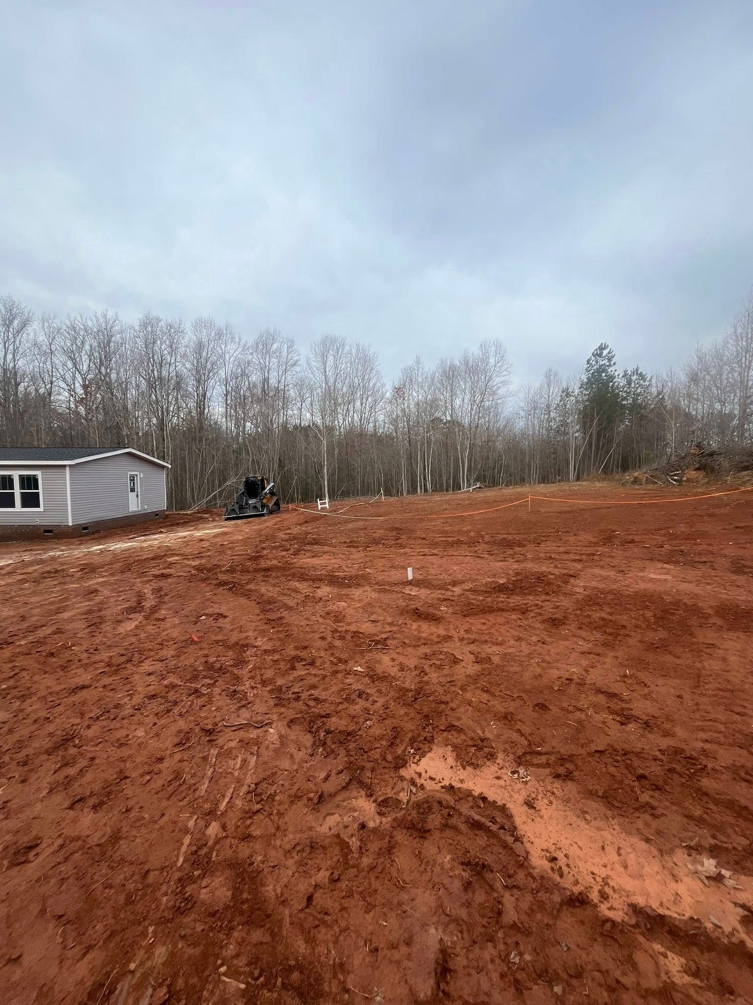 Red muddy lot with a small white house and leafless trees under an overcast sky
