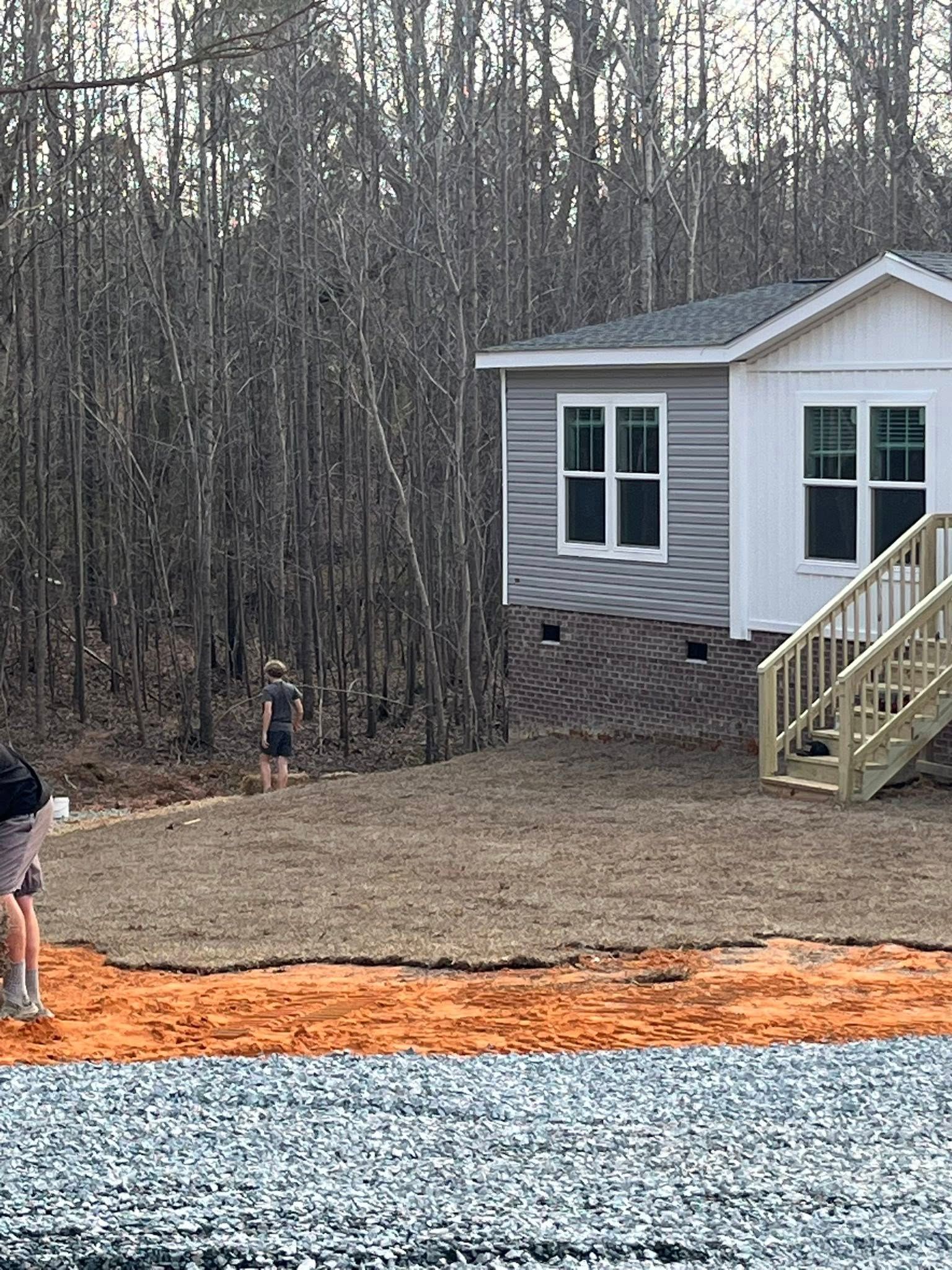 Rural yard with bare trees, a small house, and two people near a gravel driveway under construction