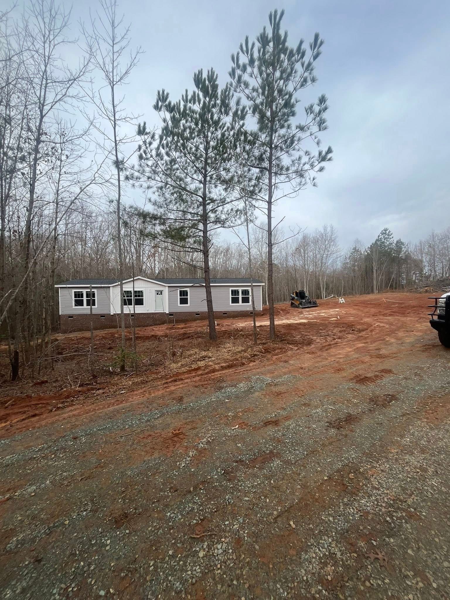 Mobile home in a wooded, muddy lot under gray skies, with a gravel road in the foreground.