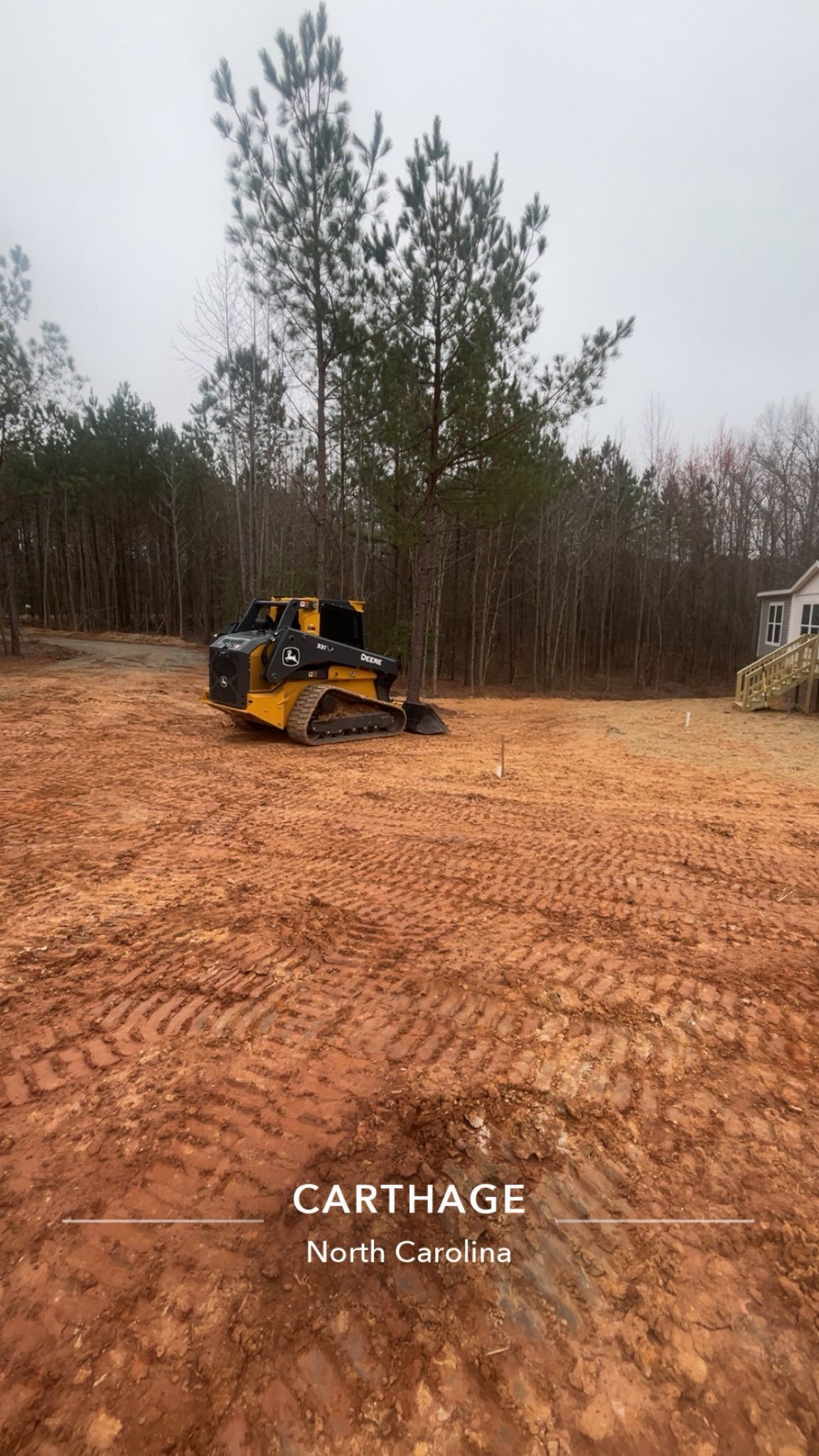 Yellow skid-steer grading a large dirt lot with trees and a house in the background