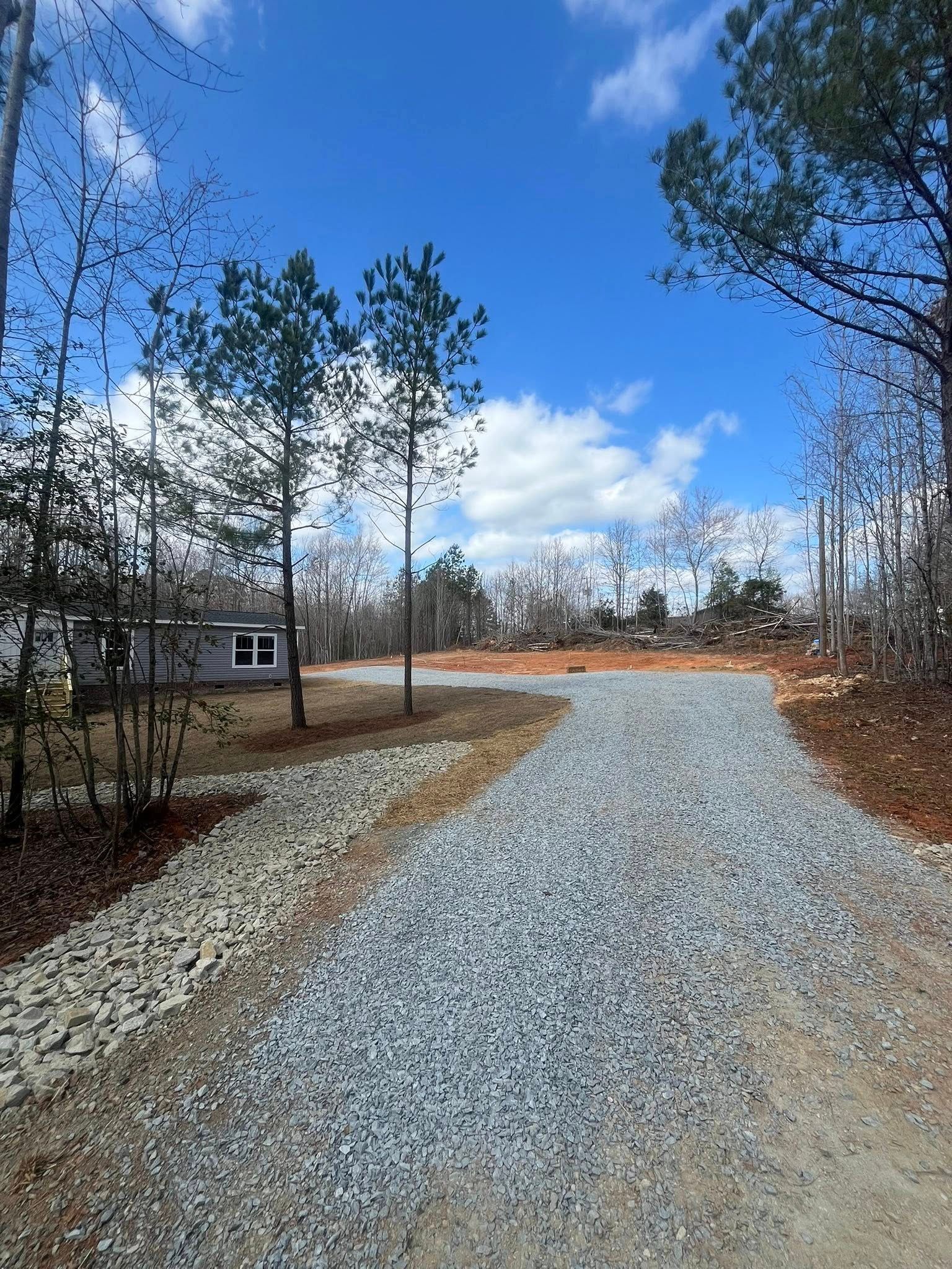 Gravel driveway curving through pine trees under a bright blue sky with clouds.