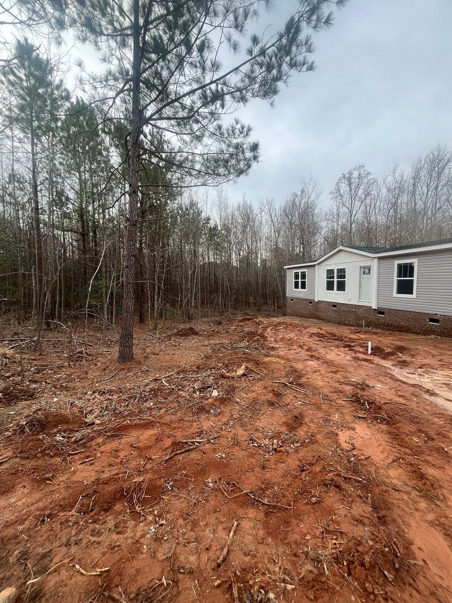 Dirt clearing beside a gray house, with trees and a cloudy sky in the background.