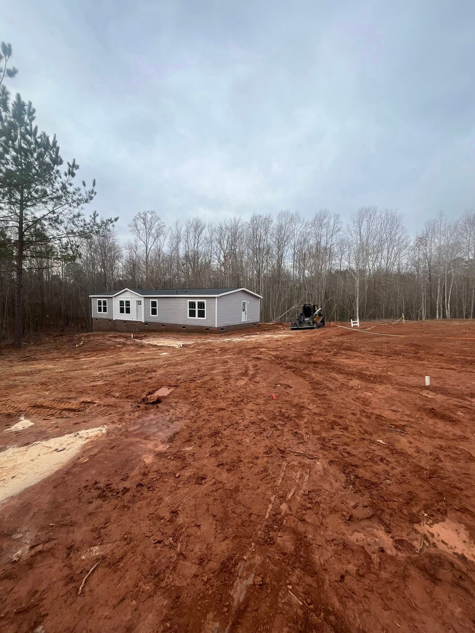 Red muddy lot leading to a white mobile home beside bare trees under a cloudy sky