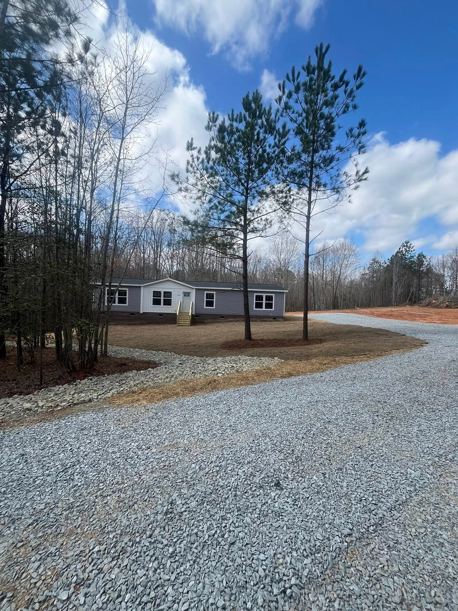 Small house beside a gravel driveway under a partly cloudy sky, surrounded by pine trees.