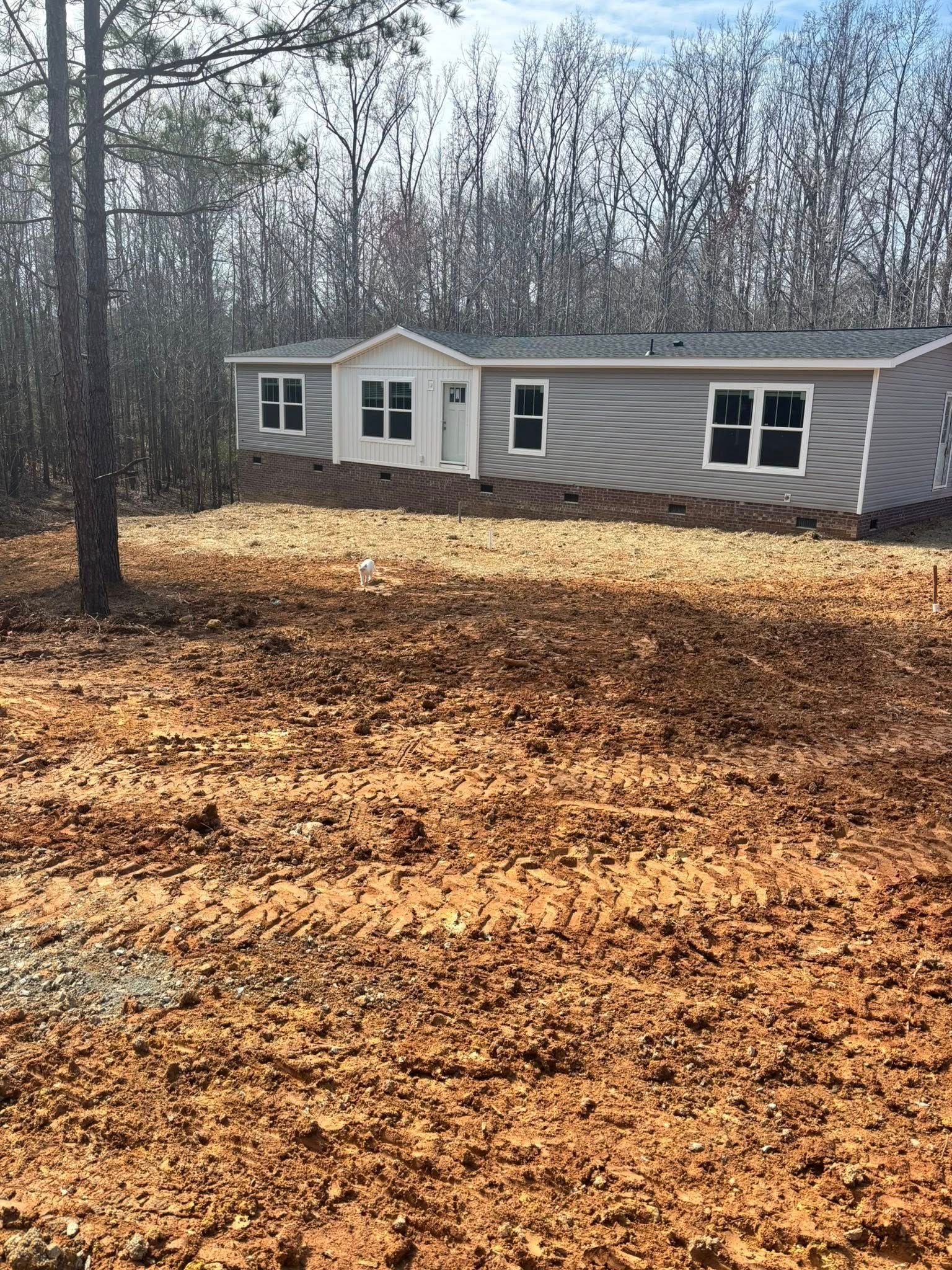 Small gray mobile home in a wooded yard covered with fallen leaves under bare trees.