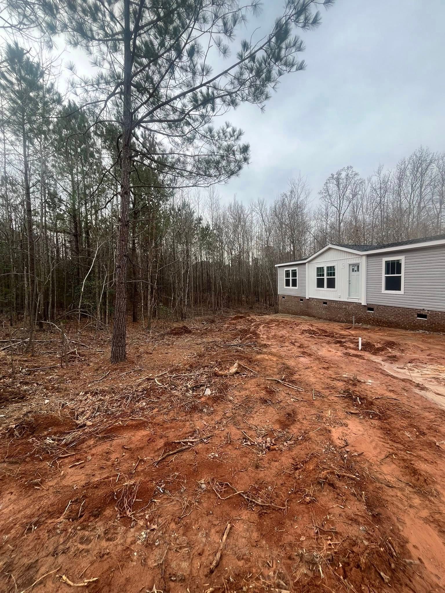 Dirt driveway beside a white house and wooded lot under a cloudy sky