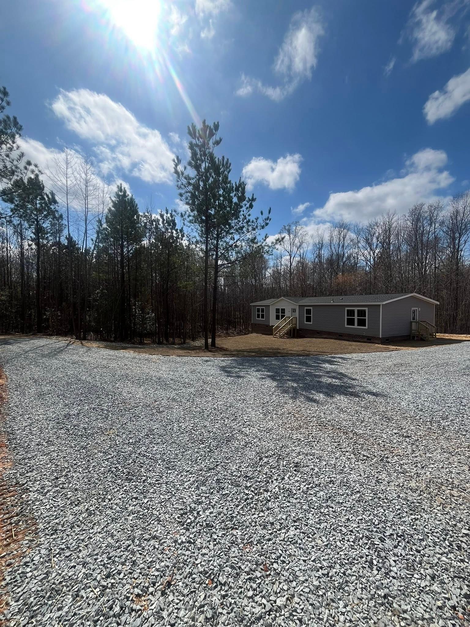 Small white house beside a gravel lot under a bright blue sky with clouds and trees