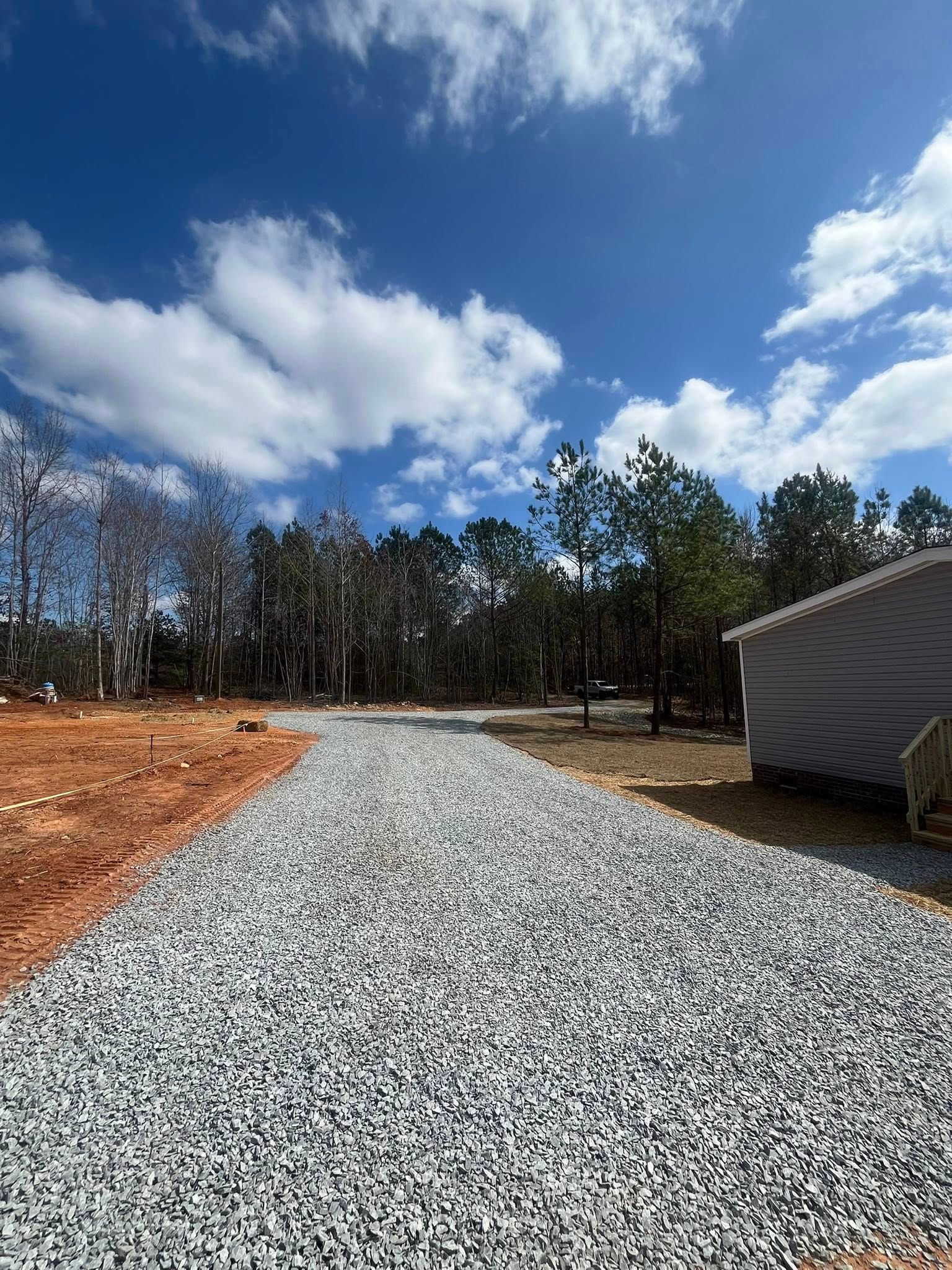 Gravel driveway leading to a wooded area under a bright blue sky with scattered clouds