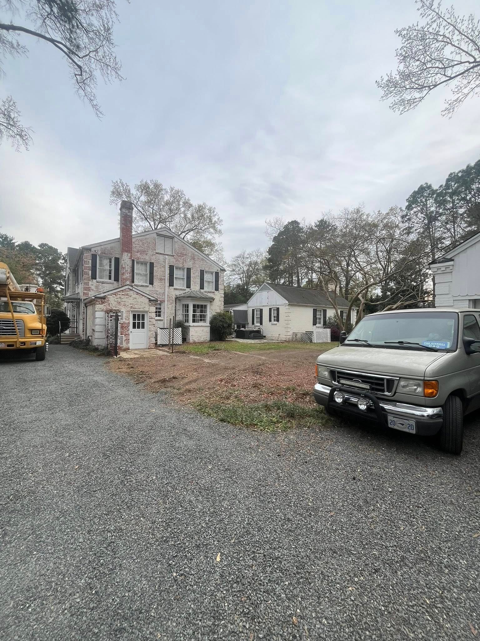 Gravel driveway with a white house, parked vehicles, and autumn trees under an overcast sky