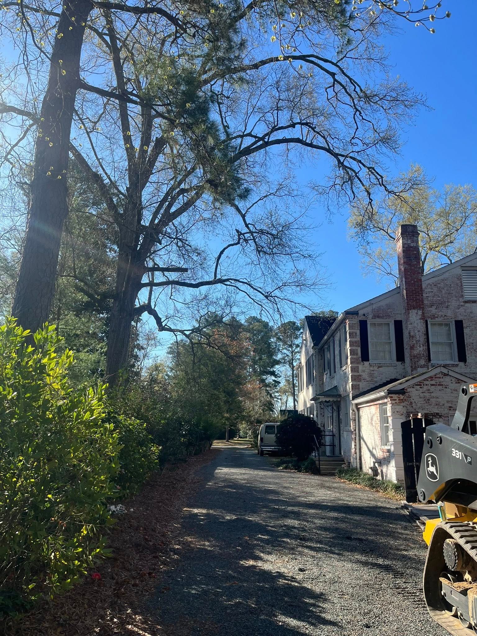 Sunny driveway beside an old white house with red chimney, trees, and parked equipment.