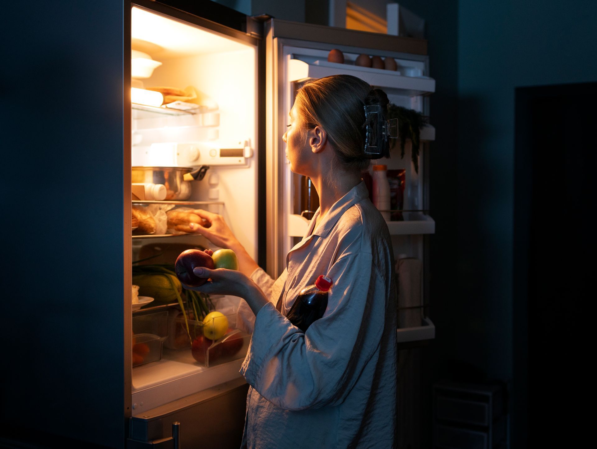 Person looking into an open refrigerator at night, holding a glass of red wine