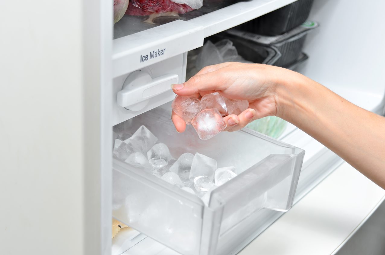 Hand reaching into an open freezer drawer to grab ice cubes from a clear bin.