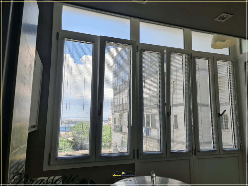 Una finestra a più vetri con vista sugli edifici e sul cielo azzurro, con cornici bianche e persiane verticali.